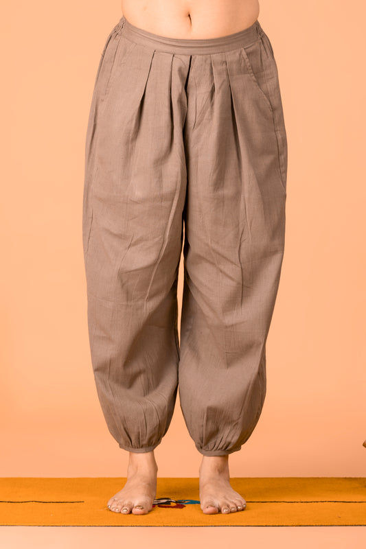 A person wearing loose, pleated harem-style pants in a taupe-brown color, standing barefoot on a yellow mat against a peach-colored background.